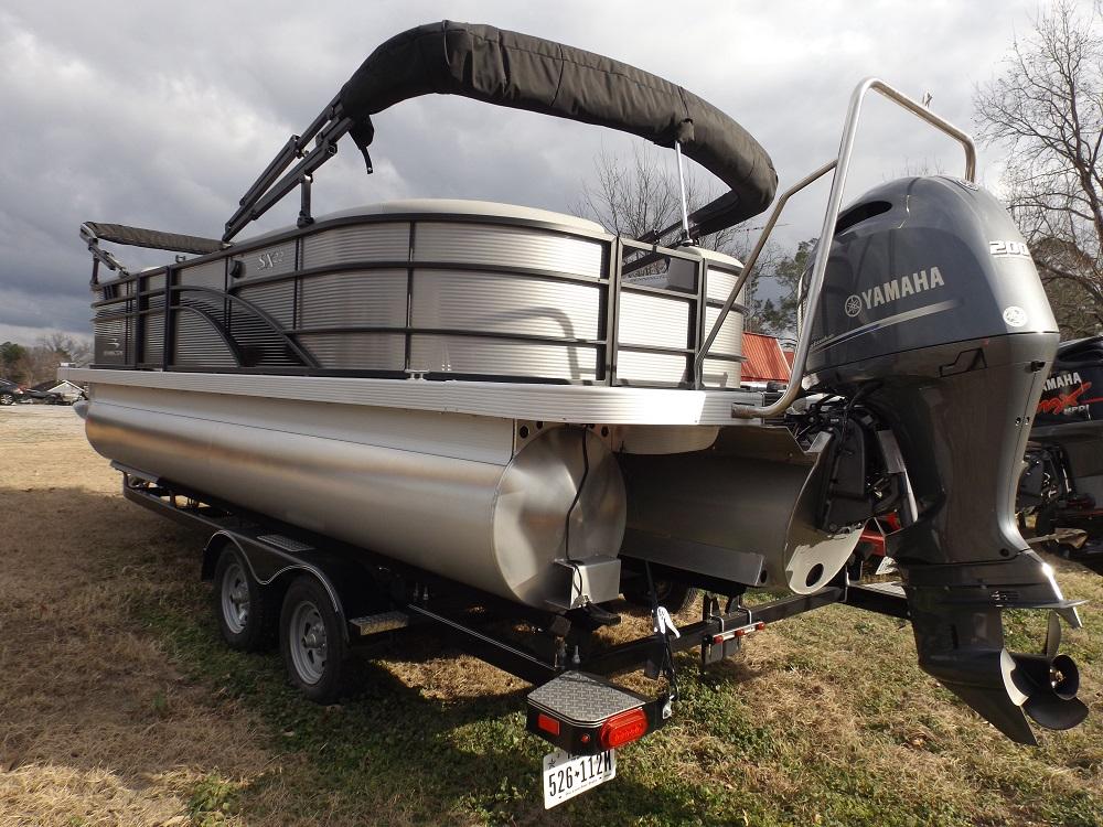 A 2023 Bennington 22 SXSR silver pontoon boat is mounted on a black trailer. The boat features an overhead canopy and is equipped with a Yamaha 200 outboard motor. The surroundings appear to be an outdoor area with grass and trees under a cloudy sky.