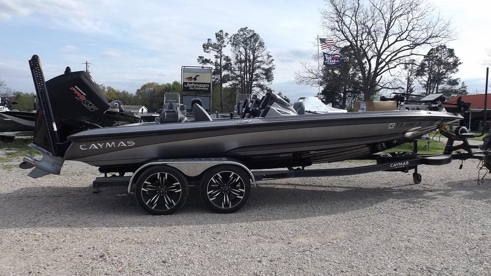 A sleek gray 2021 Caymas CX21Pro bass fishing boat is parked on a gravel lot, attached to a double-axle trailer with black and silver wheels. Various trees and a few flags can be seen in the background.