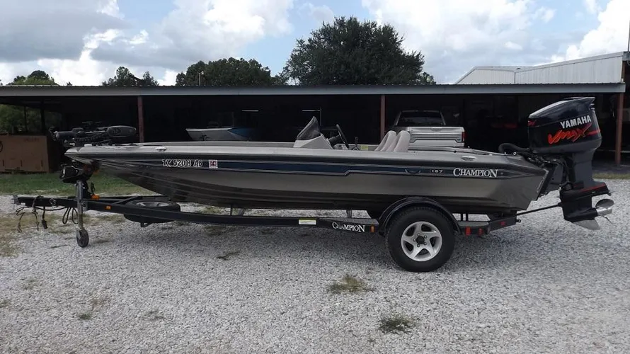 Alt text: "19-foot Champion 187 fishing boat with Yamaha outboard motor on trailer at Lake Fork, parked on gravel surface near building and trees. Daytime with partly cloudy skies.