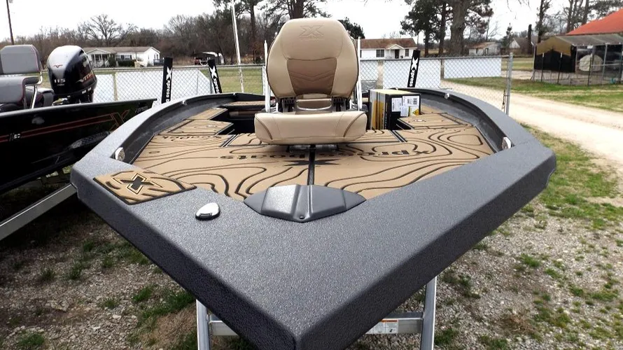 2024 Xpress H20 fishing boat front deck with tan cushioned chair, storage compartments, and patterned tan surface on a trailer in Lake Fork. Grass, trees, and houses in the background showcase the outdoor setting.