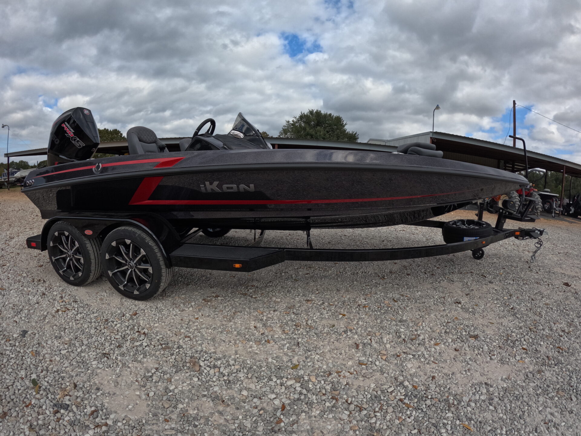 2025 iKon VLX21 motorboat in black and red on trailer at Sartin Marine, Lake Fork, showcasing modern design and prominent motor against lush trees and cloudy sky.