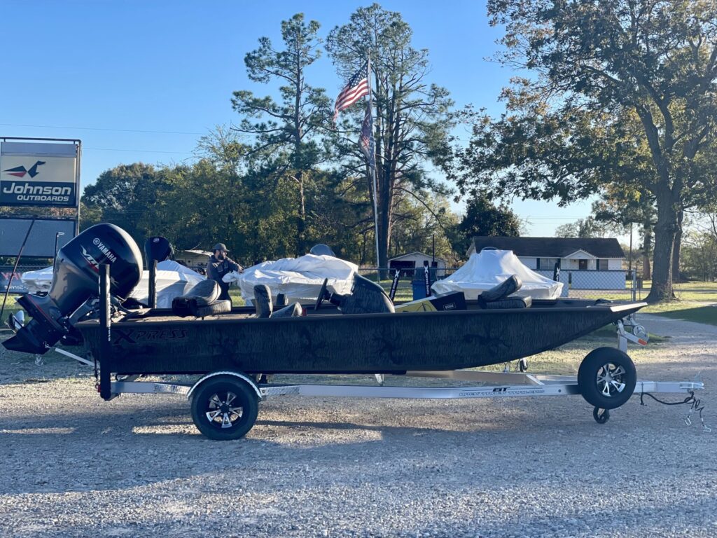 Black Xpress HP180 CAMO fishing boat with Yamaha 90 motor on trailer at Sartin Marine, Lake Fork. American flag near Johnson Outdoors sign, surrounded by covered boats and framed by trees under a clear sky. Serene 2025 aquatic scene.