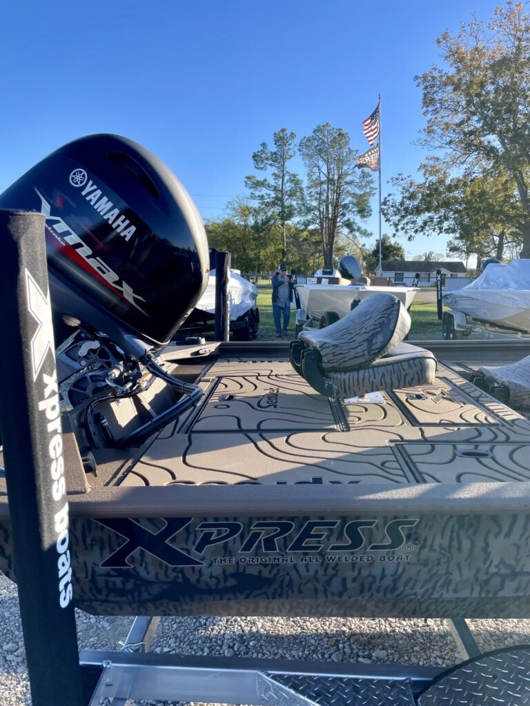 Camo Xpress HP180 fishing boat with Yamaha 90 motor on trailer at Sartin Marine, Lake Fork. Features seats and fishing gear, "Xpress Boats" logo visible. American flag and blue sky in background.