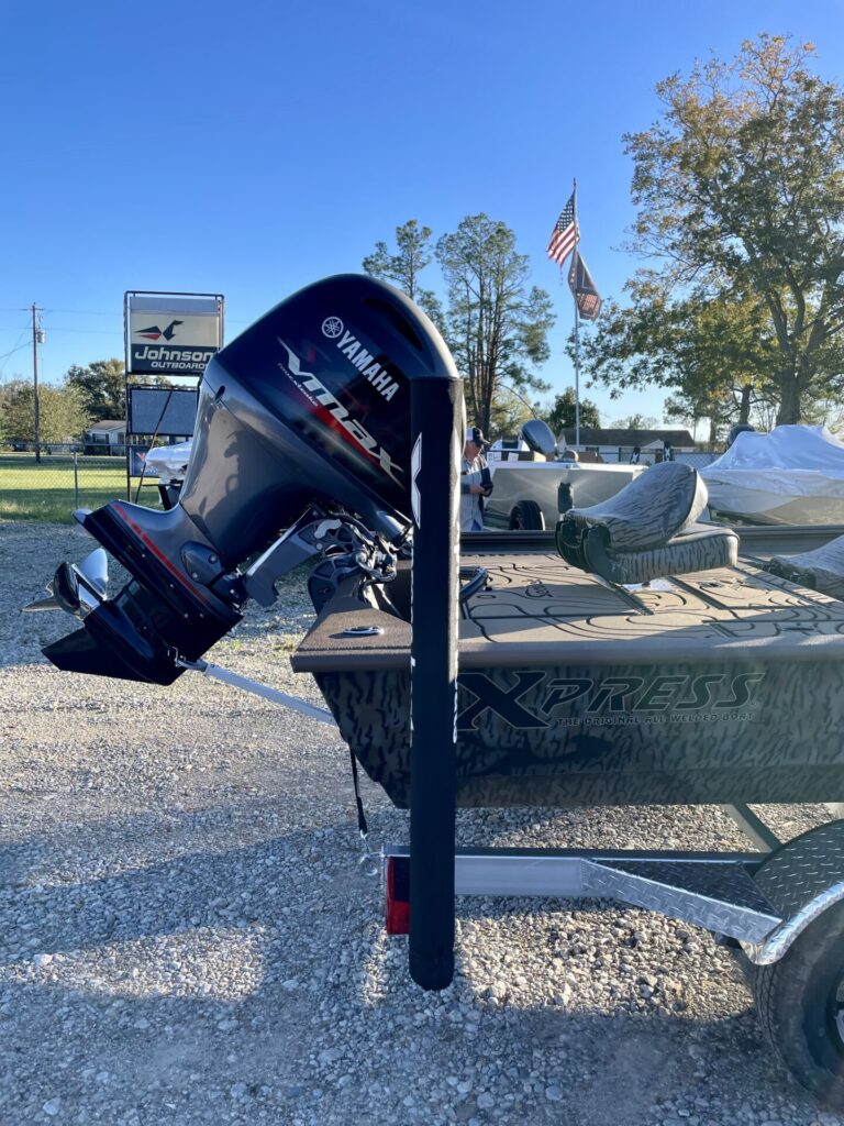 Alt text: "XPRESS HP180 CAMO boat with Yamaha 90 motor on trailer at Sartin Marine, Lake Fork. Johnson sign, trees, and American flags in sunny 2025 sky.
