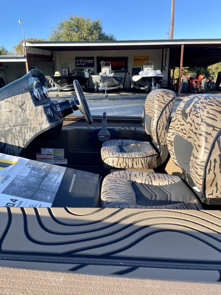 Image of Sartin Marine at Lake Fork showcasing the interior of a BASS HP180 boat with camouflage-patterned seats and dashboard. The prominent steering wheel and control lever are highlighted, while several boats, including the Xpress Xplorer, are parked under a metal shelter. Sunlight creates artistic shadows across the scene, emphasizing marine equipment ready for customers.
