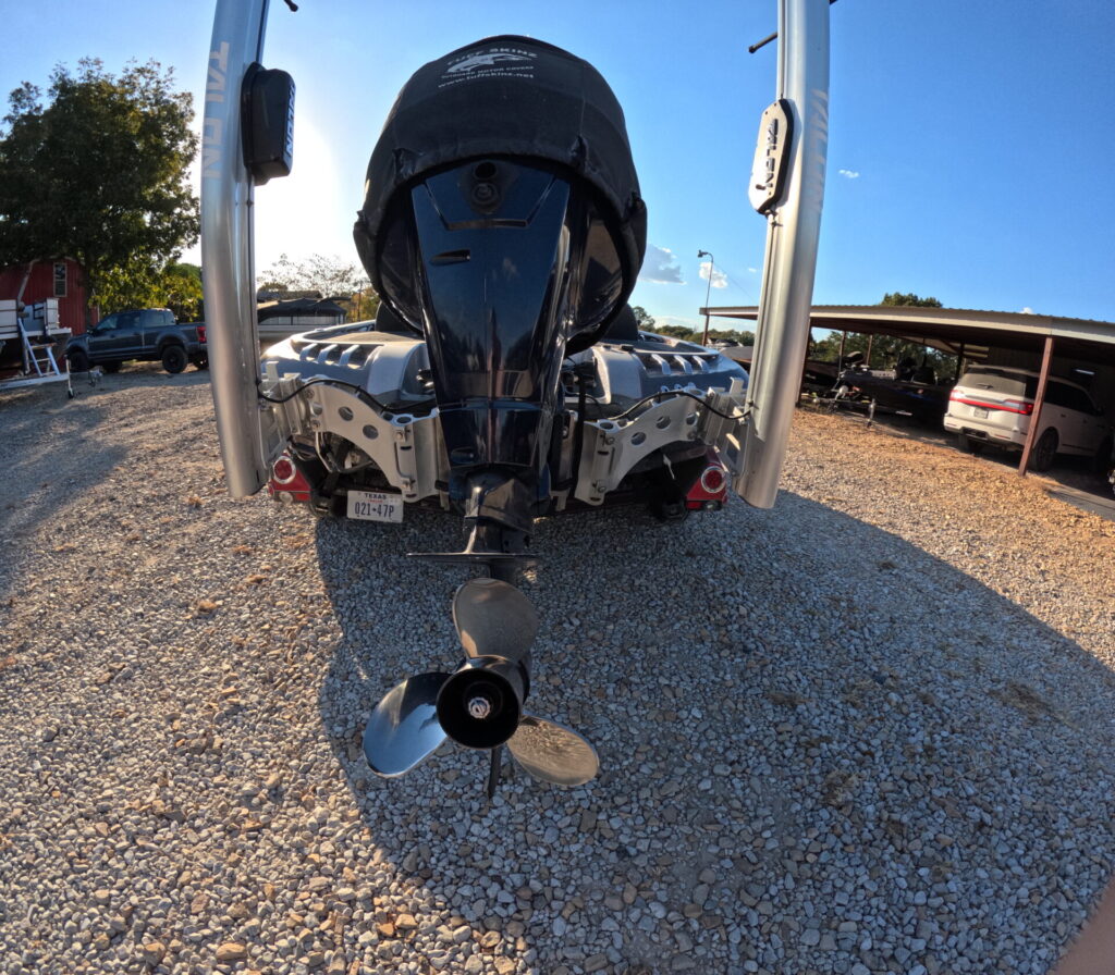 Alt text: "2012 Ranger Comanche Z520S boat showcasing motor and propeller on gravel at Sartin Marine, Lake Fork; buildings and vehicles in the background beneath a clear blue sky.