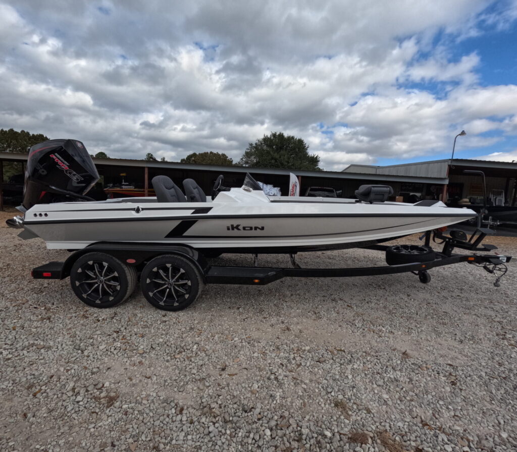 White and black iKon fishing boat on a black trailer in a gravel lot at Lake Fork. Features sleek hull, multiple seating areas, and outboard motor. Overcast sky with buildings in the background, showcasing modern marine equipment at Sartin Marine.